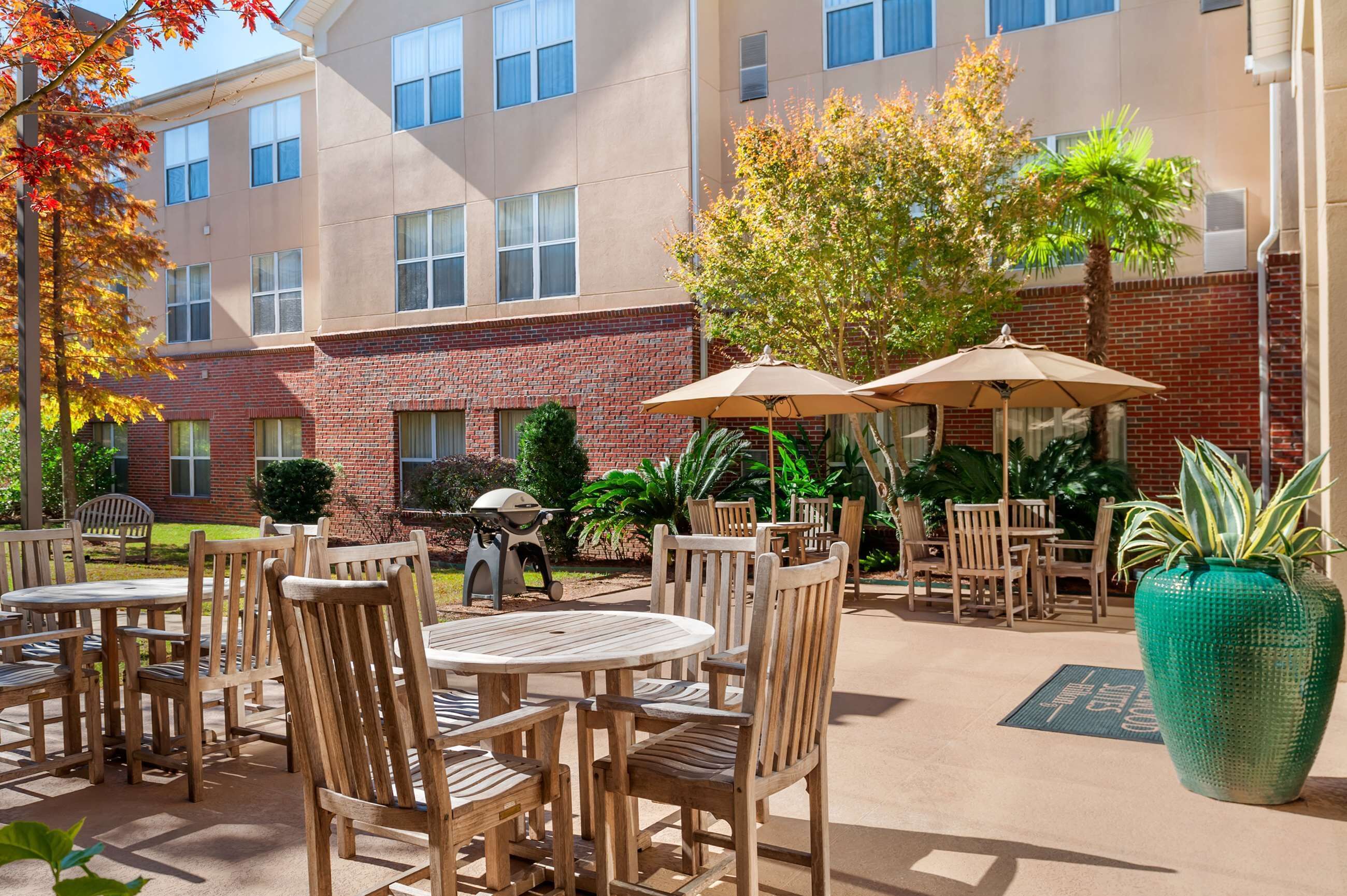 An outdoor patio area with wooden tables and chairs, umbrellas, and a large green planter.