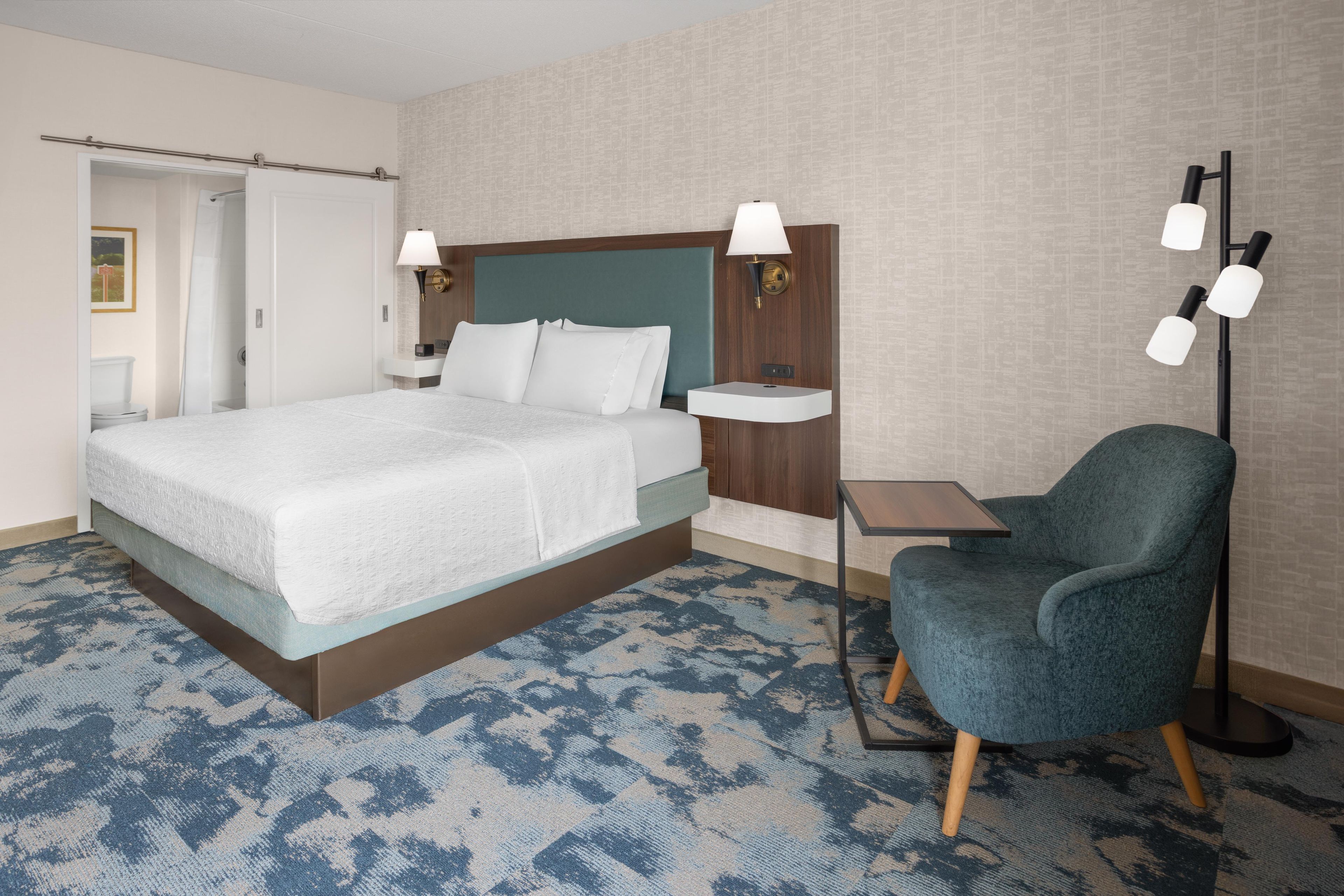 A hotel room with a bed, bedside table, chair, and a floor lamp.
