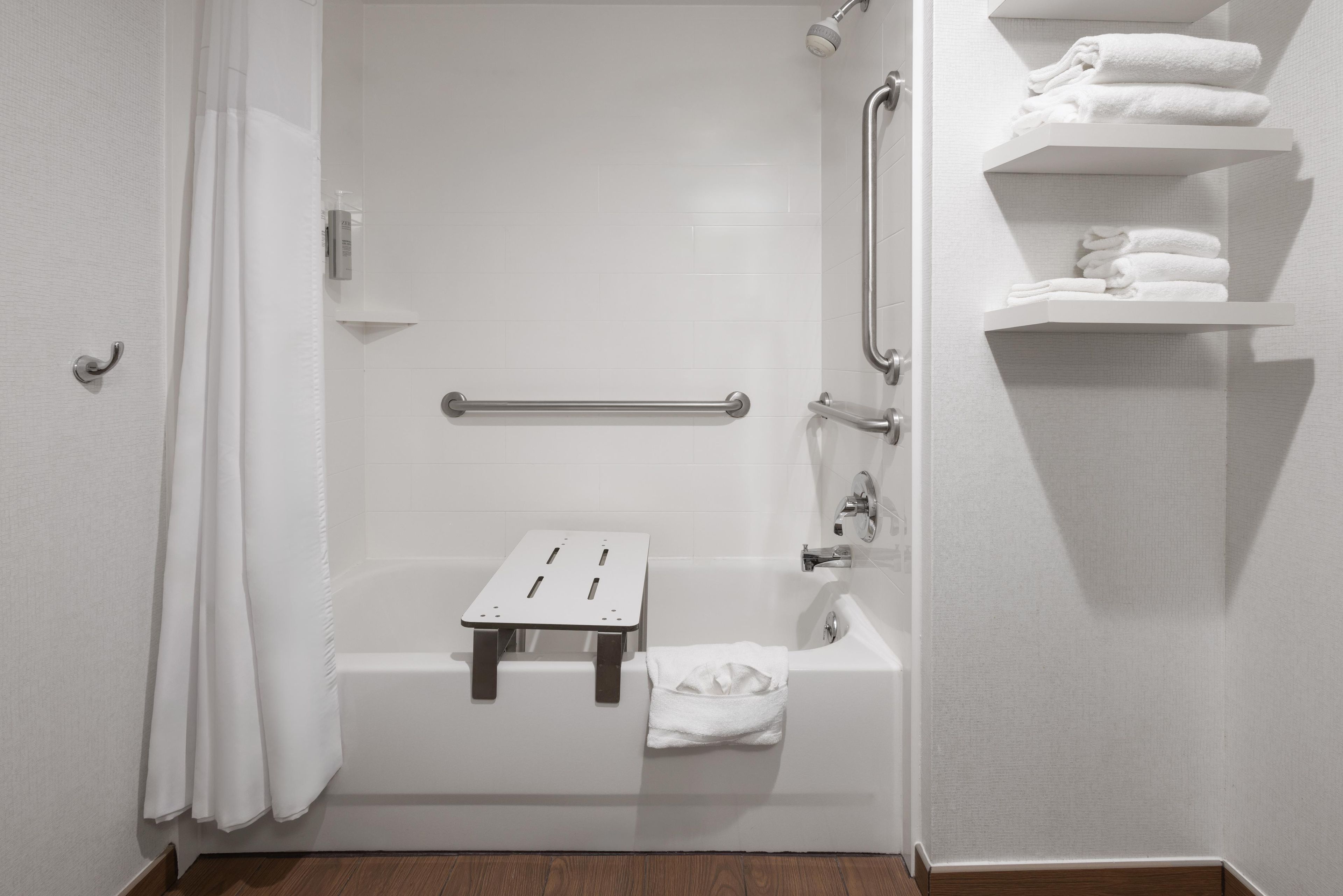 A bathroom with a white bathtub, a shower, a grab bar, a towel rack, and a shelf with folded towels.