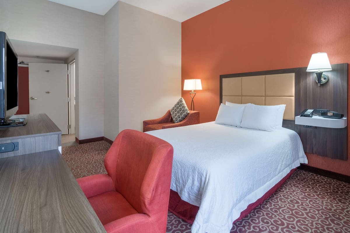 A hotel room with a bed, a desk with a television, a chair, a lamp, and a wall-mounted phone.