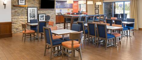 A hotel dining area with multiple tables and chairs, a stone wall with framed pictures, and a counter with a menu.