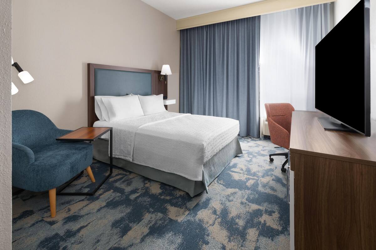 Une chambre d’hôtel équipée d’un lit, d’un bureau, d’une chaise, d’une télévision et d’une grande fenêtre avec des rideaux.
