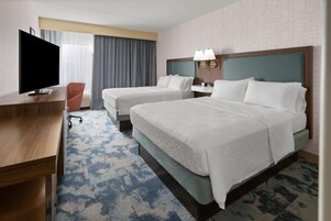 A hotel room with a flat-screen TV, two beds with white linens, a desk, a chair, a nightstand with lamps, and a patterned carpet.