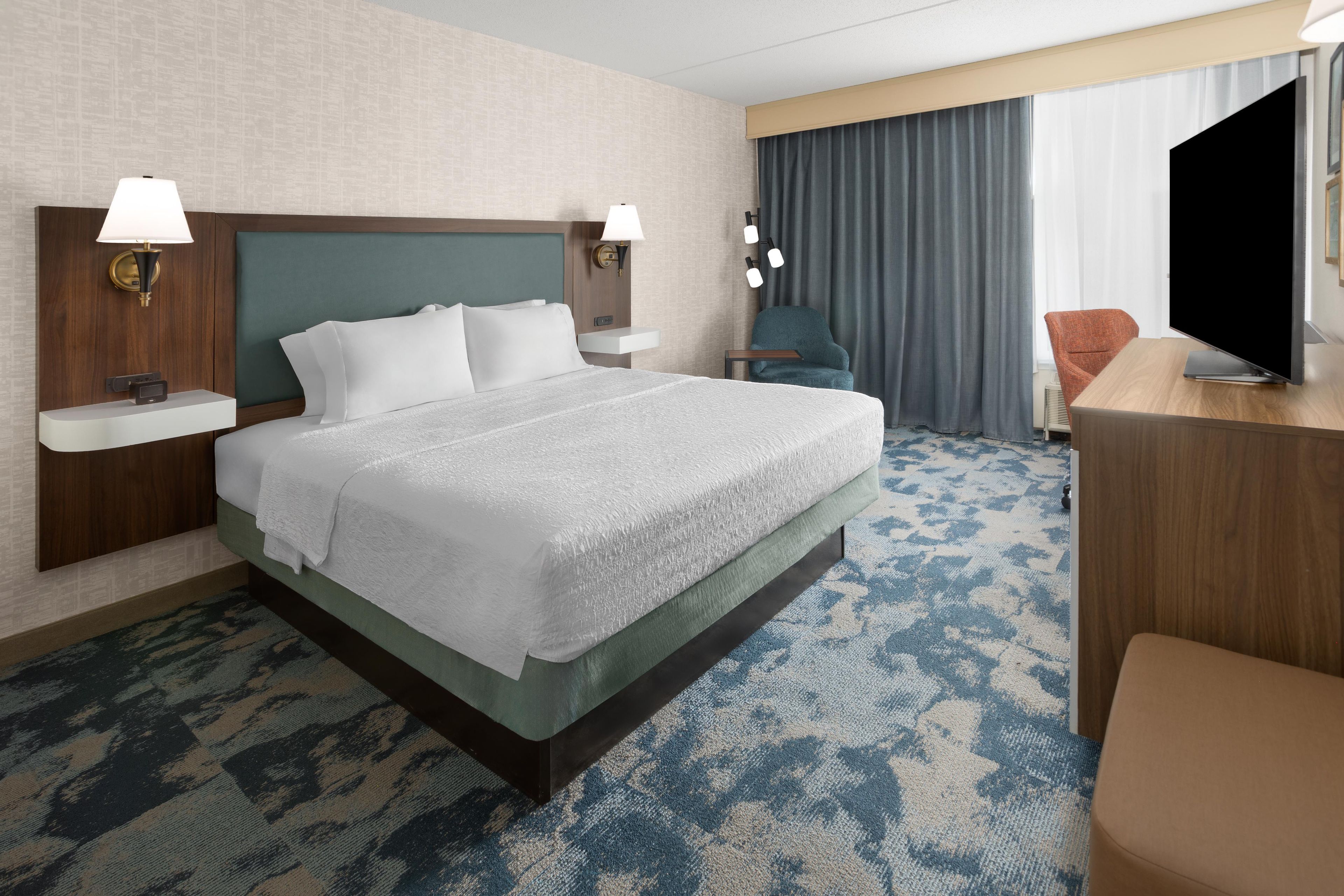 Una habitación de hotel con una cama grande, dos mesitas de noche, un escritorio con silla, un televisor y una ventana con cortinas.