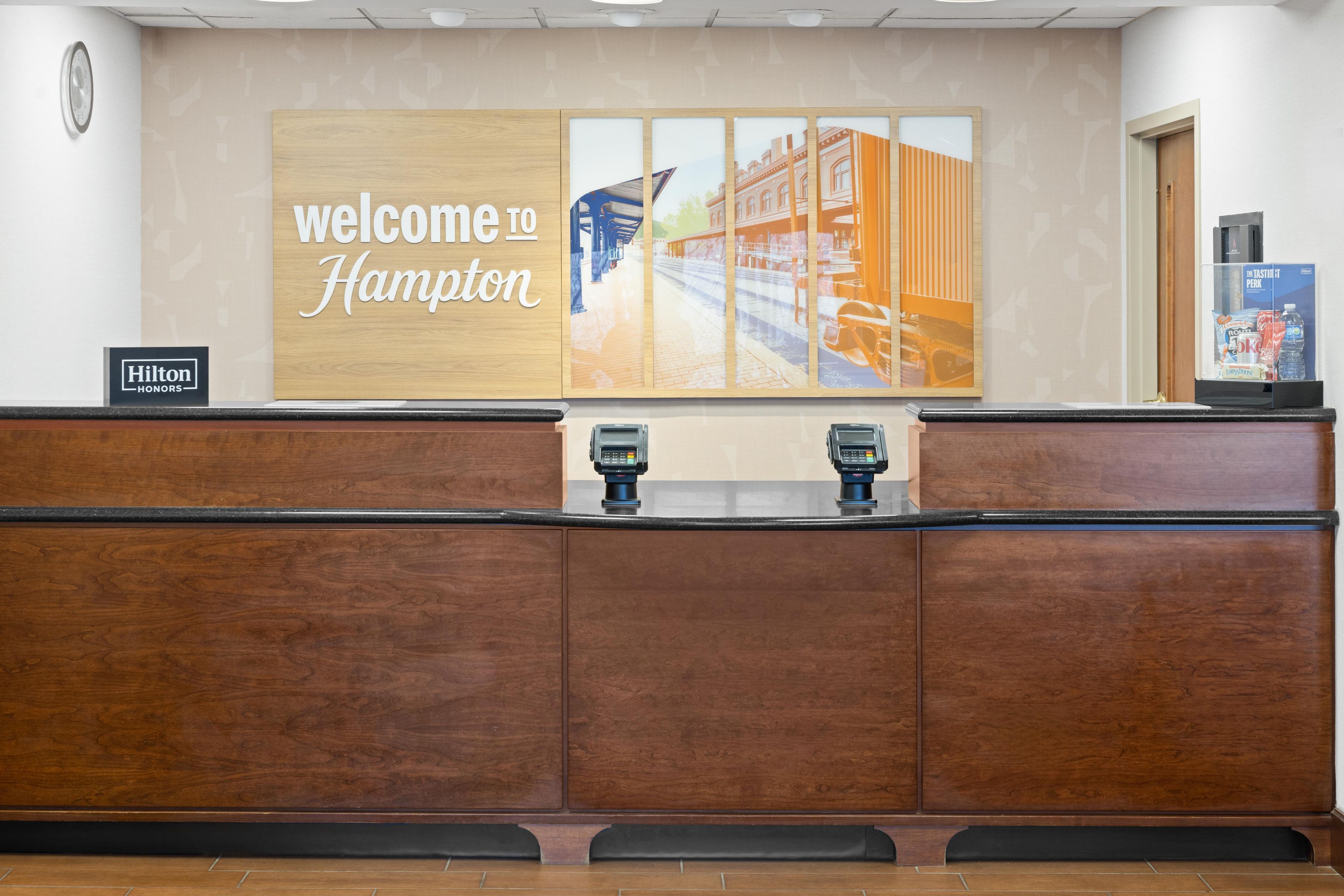A reception desk with a 'Welcome to Hampton' sign, a clock, and a minibar.