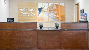 A reception desk with a 'Welcome to Hampton' sign, a clock, and a minibar.