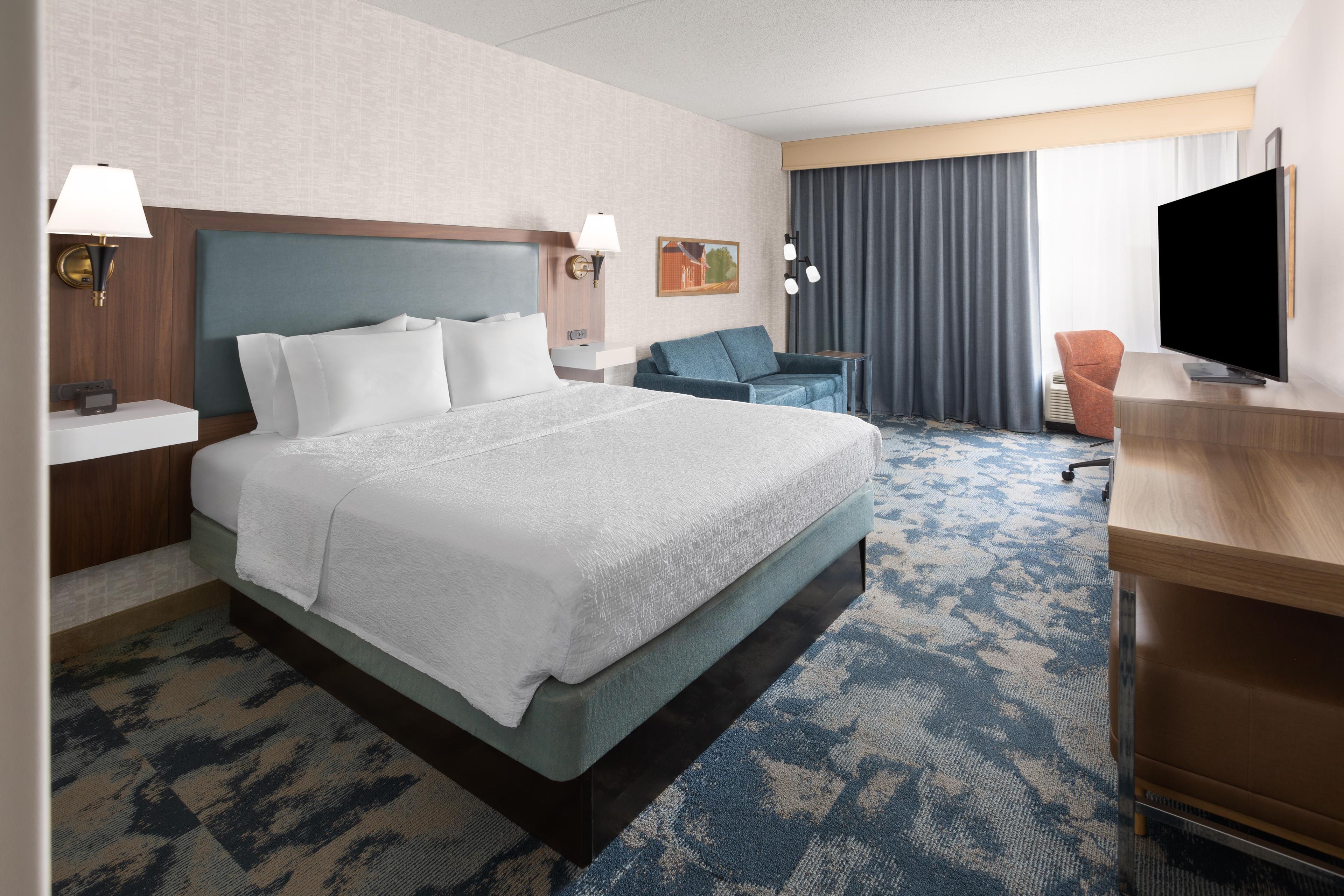 A hotel room with a large bed, a desk, a chair, a television, and a window with curtains.
