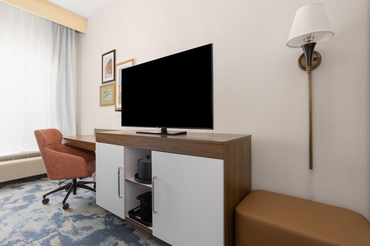 Une chambre d’hôtel avec un bureau en bois, une télévision, un banc rembourré, une lampe fixée au mur et des tableaux encadrés accrochés au mur.