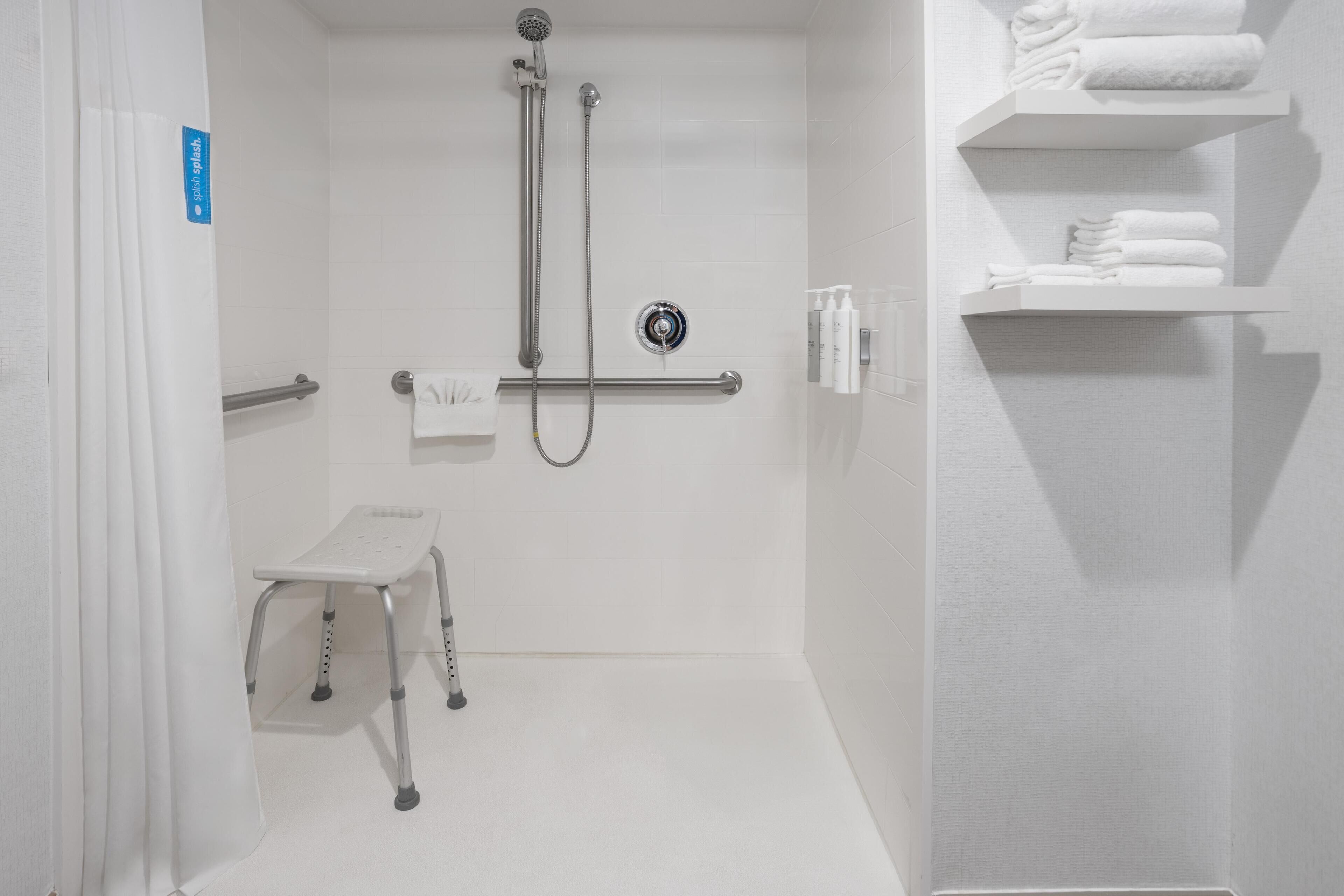 A white bathroom with a shower, a grab bar, a small stool, and shelves with towels.