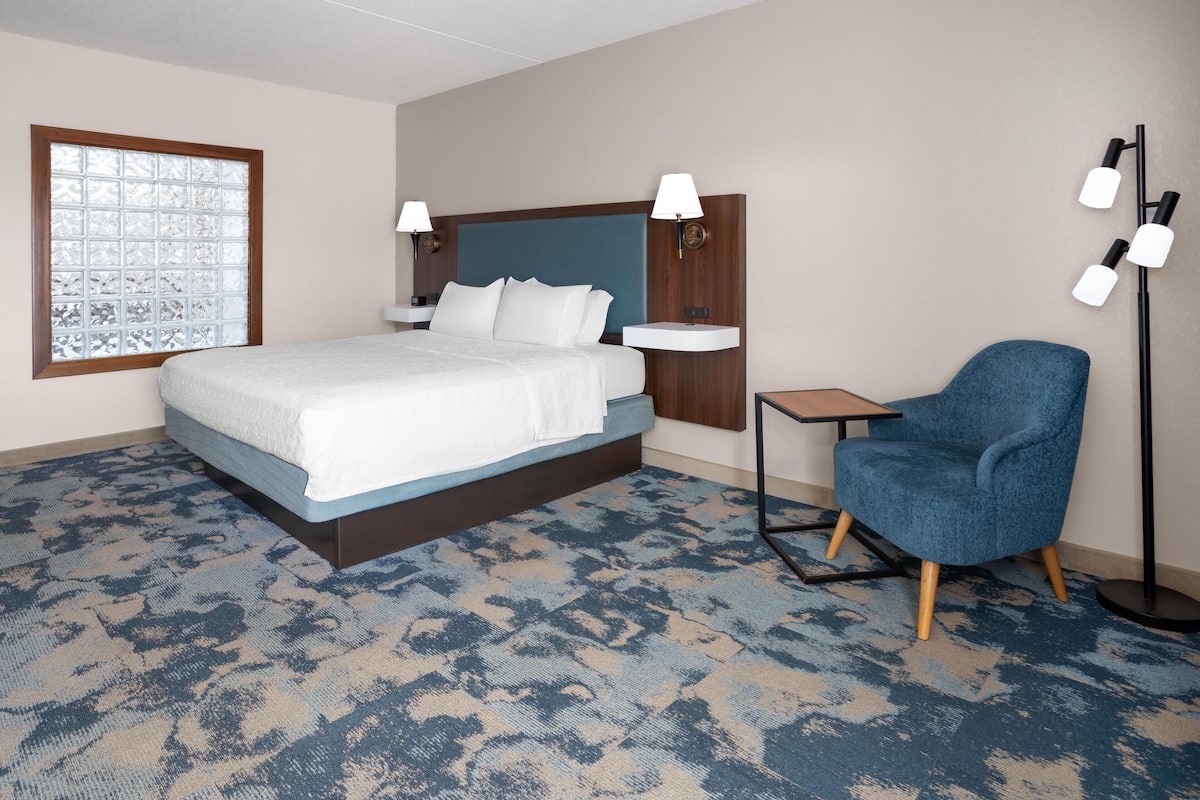 Une chambre d’hôtel avec un lit, une chaise, une table de chevet, une lampe et une fenêtre à vitre dépolie.