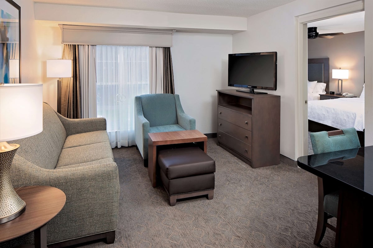 Een hotelkamer met een bank, fauteuil, poef, televisie, dressoir en een bed met nachtkastje.