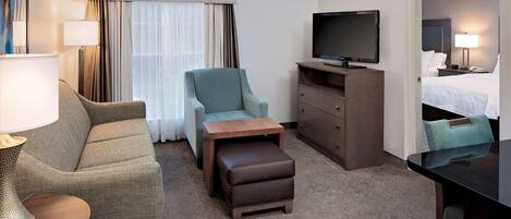 Habitación de hotel con sofá, sillón, reposapiés, televisión, cómoda y cama con mesita de noche.