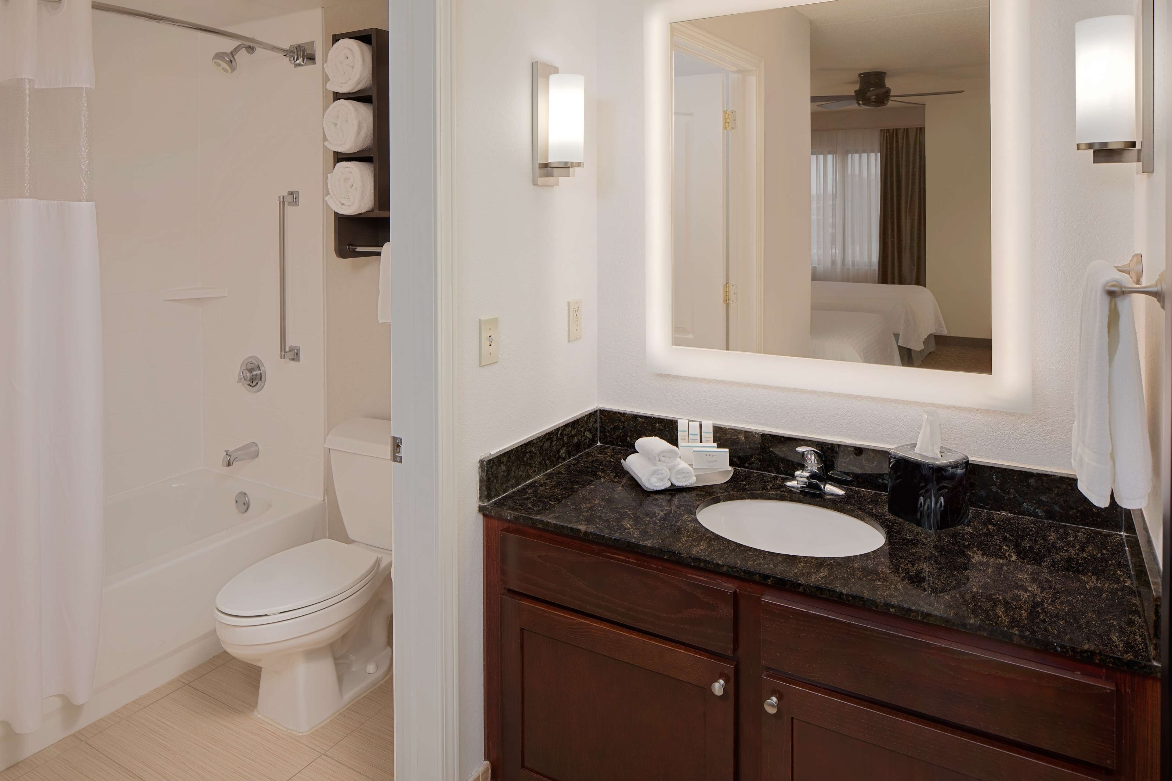 Une salle de bain avec des toilettes blanches, une douche, un lavabo doté d’un plan de toilette foncé et un miroir au-dessus du lavabo.
