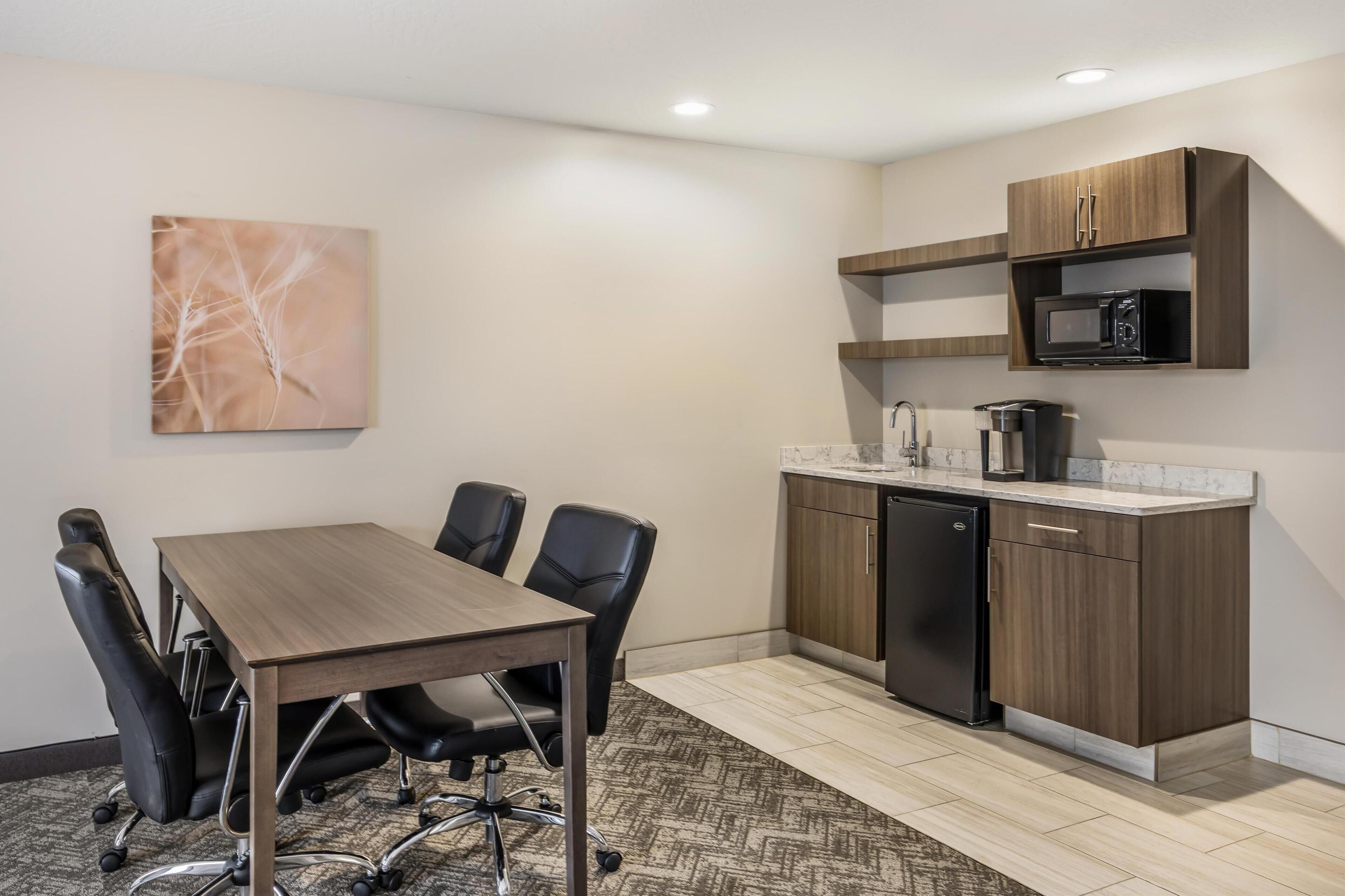 A conference room with a wooden table and black chairs, a kitchen with a microwave and dishwasher, and a wall-mounted shelf.