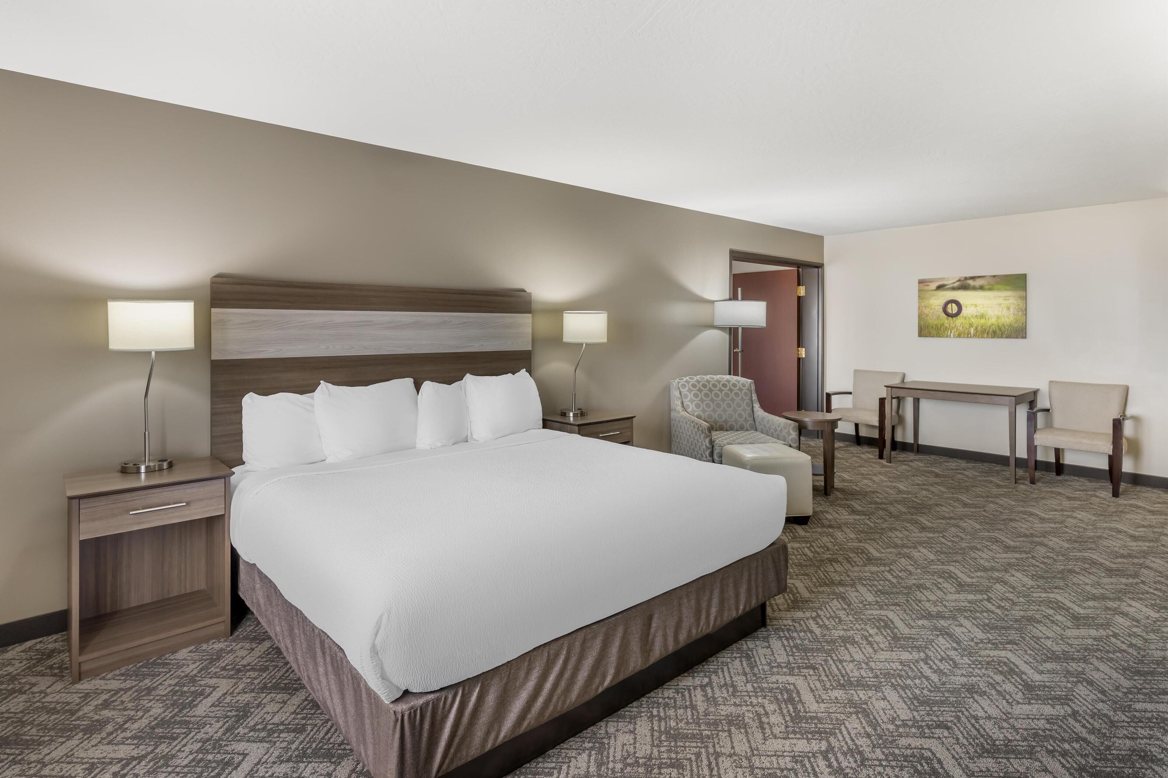 A hotel room with a large bed, two bedside tables with lamps, a desk with two chairs, and a painting on the wall.