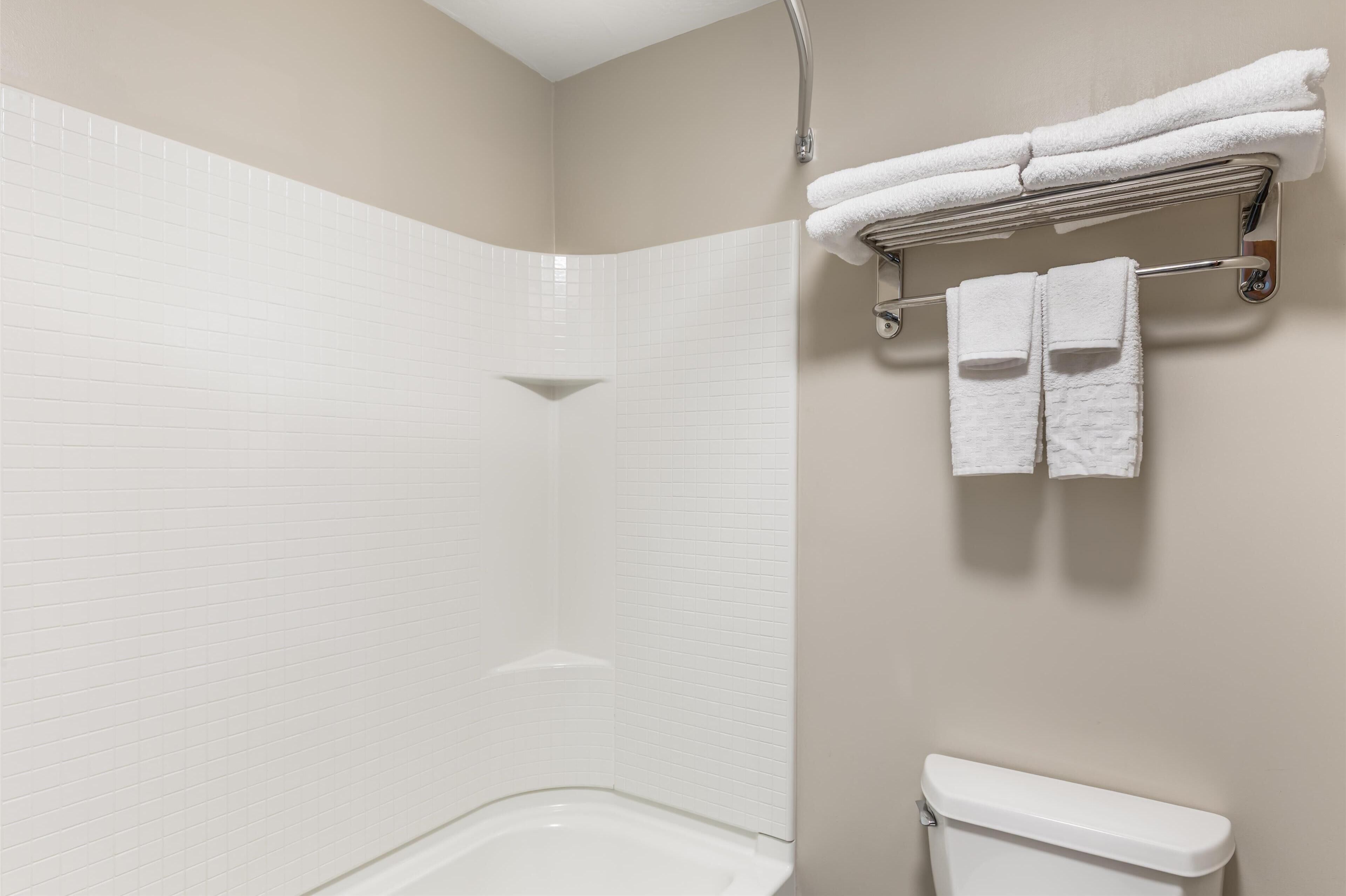 Une salle de bain avec des murs recouverts de carreaux blancs, une baignoire blanche et des toilettes. Deux serviettes blanches sont suspendues à un porte-serviettes en métal.
