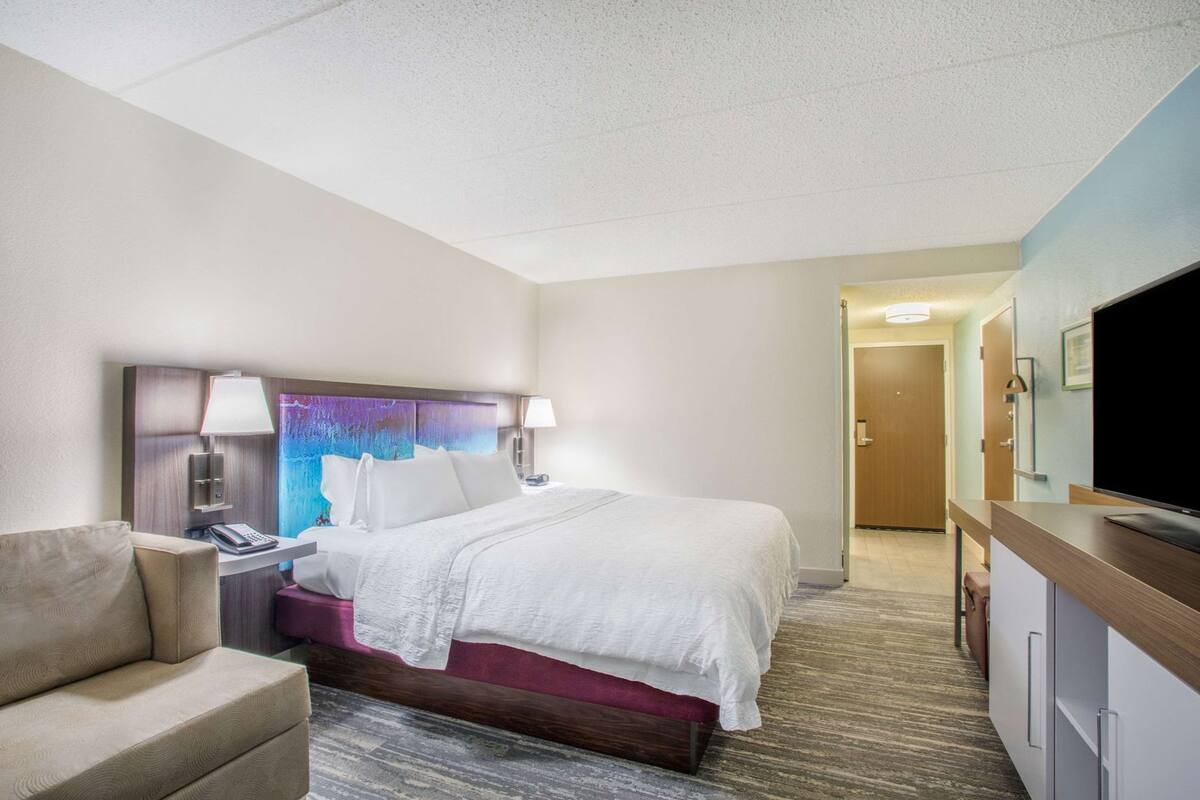 A hotel room with a bed, bedside tables, a chair, a television, and a door leading to another room.