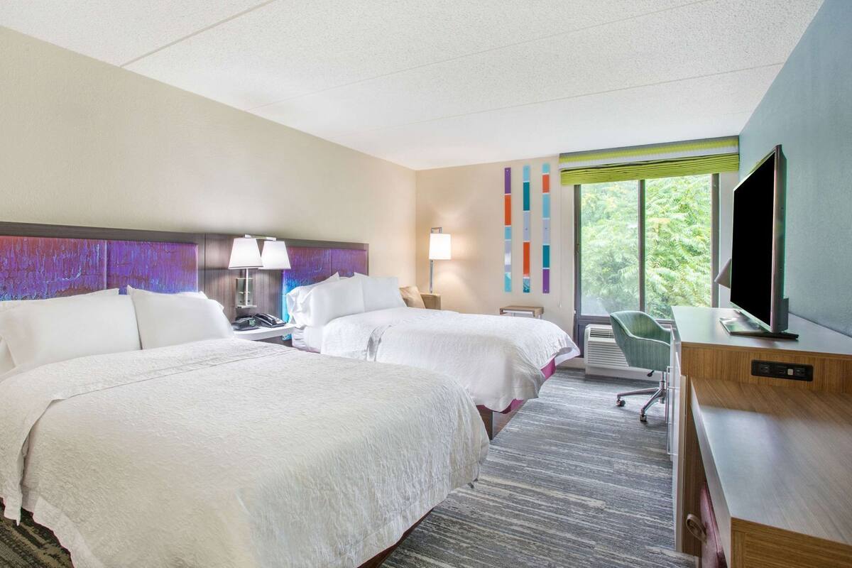 A hotel room with two beds, a desk with a TV, a chair, and a window with a view of trees.