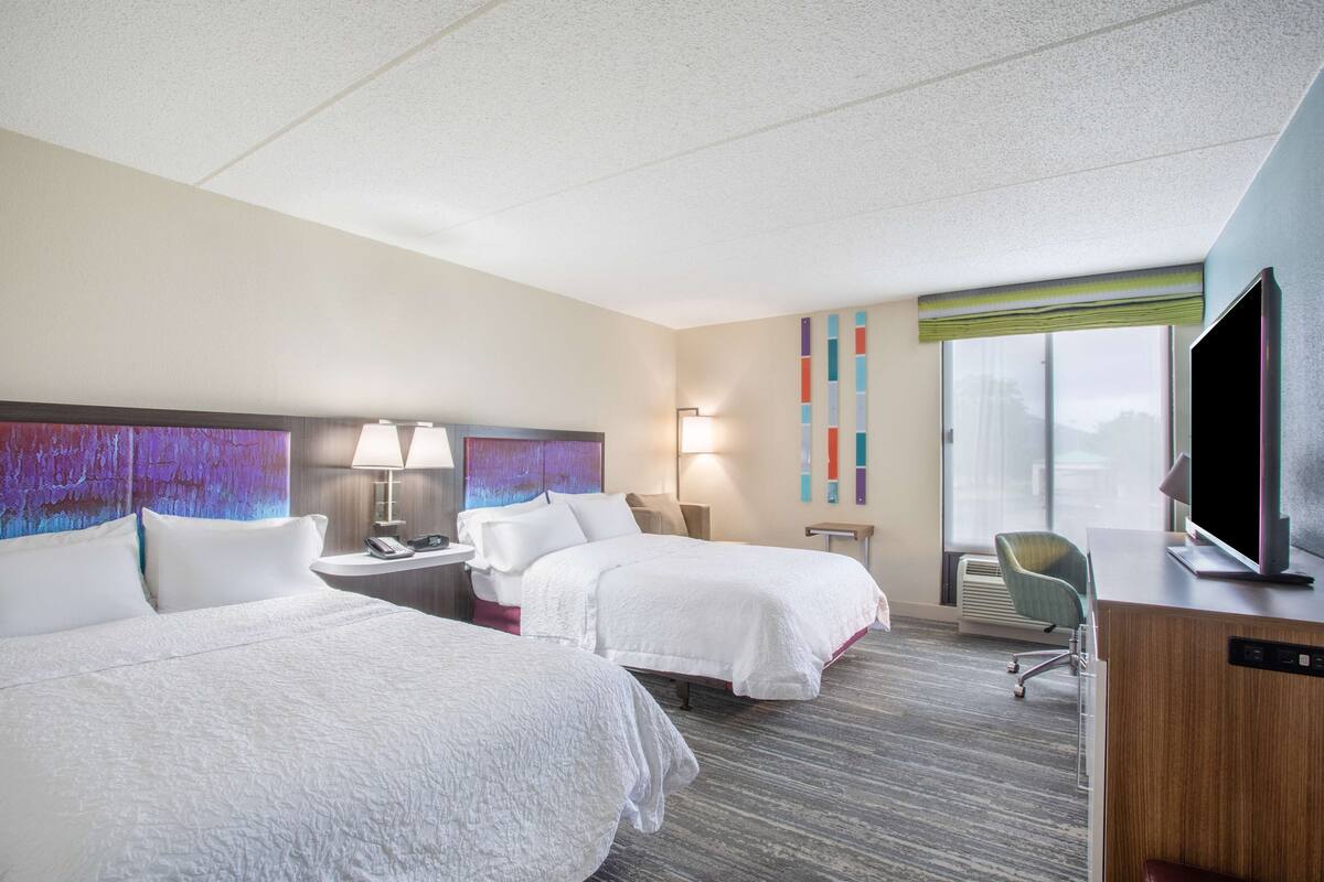 A hotel room with two beds, a desk, a television, and a window with blinds.