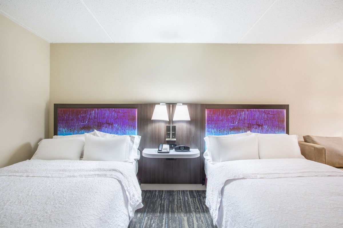 A hotel room with two beds, a small desk with a lamp, and a wall-mounted phone.