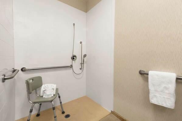 A bathroom with white tiled walls, a beige floor, a grab bar, a wheelchair, a shower with a handheld showerhead, and a towel rack.