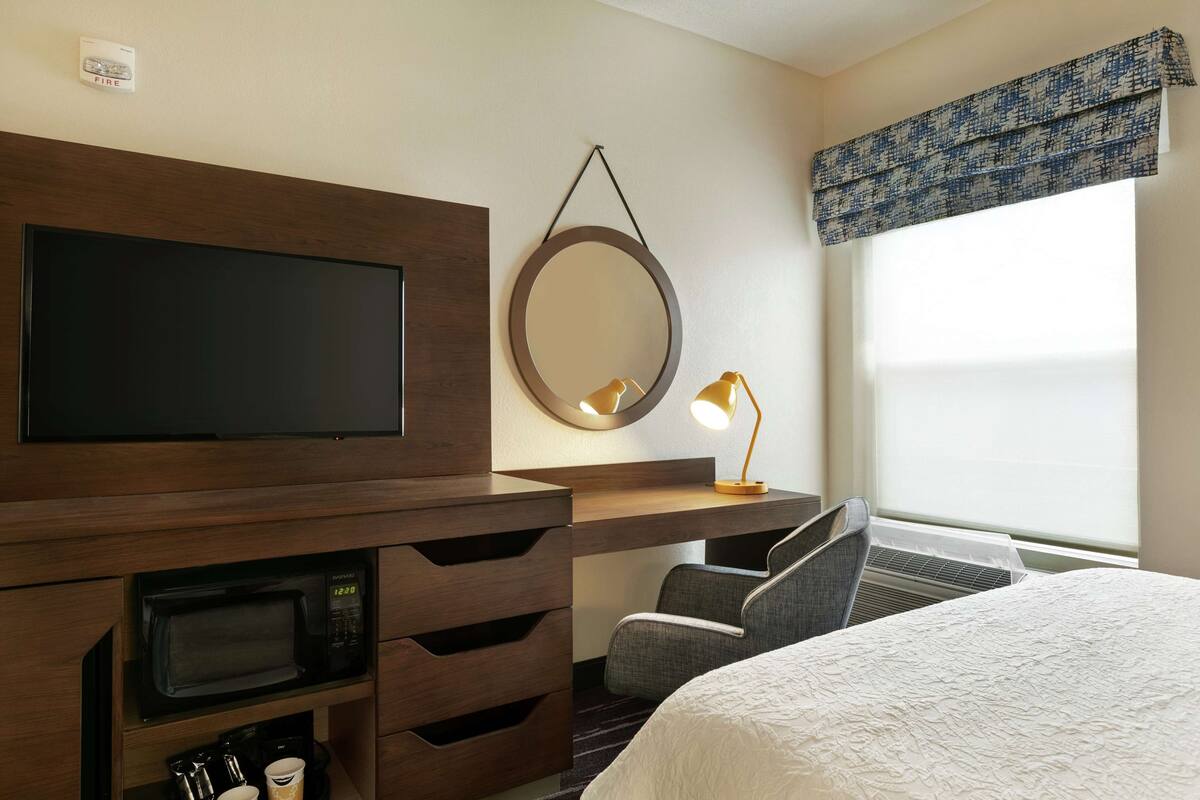 A hotel room with a flat-screen TV, a microwave, a desk with a lamp, a chair, a bed, and a large window with a view.