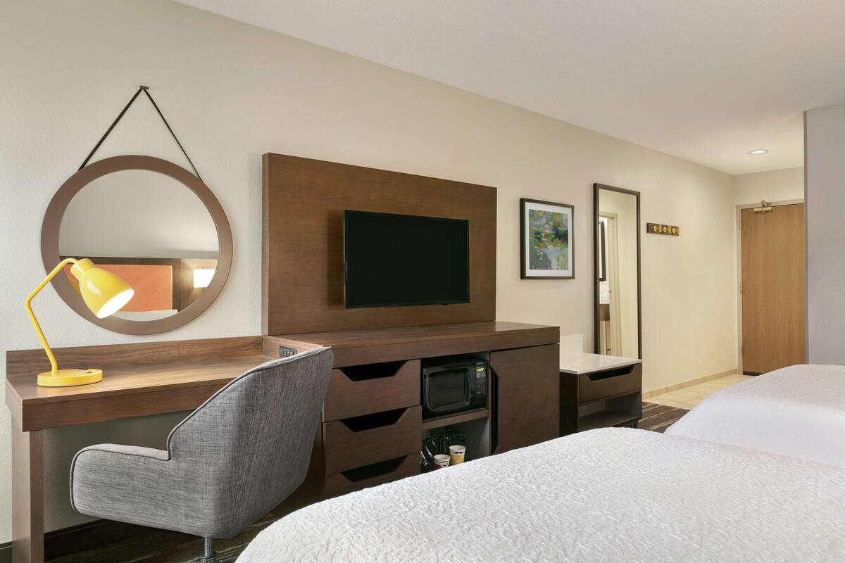 A hotel room with a desk, a chair, a TV, a microwave, a bed, and a mirror.