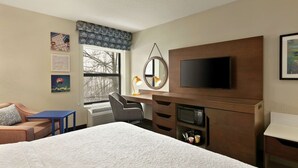 Ein Hotelzimmer mit Bett, Schreibtisch mit Computer, Fernseher und Fenster mit Blick auf Bäume.