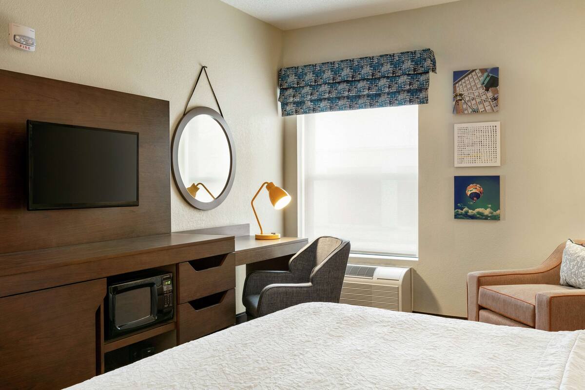 A hotel room with a bed, a desk with a chair, a television, a microwave, and a window with a view of buildings.