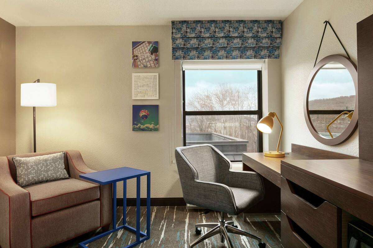A hotel room with a sofa, desk, chair, lamp, and a large window with a view.