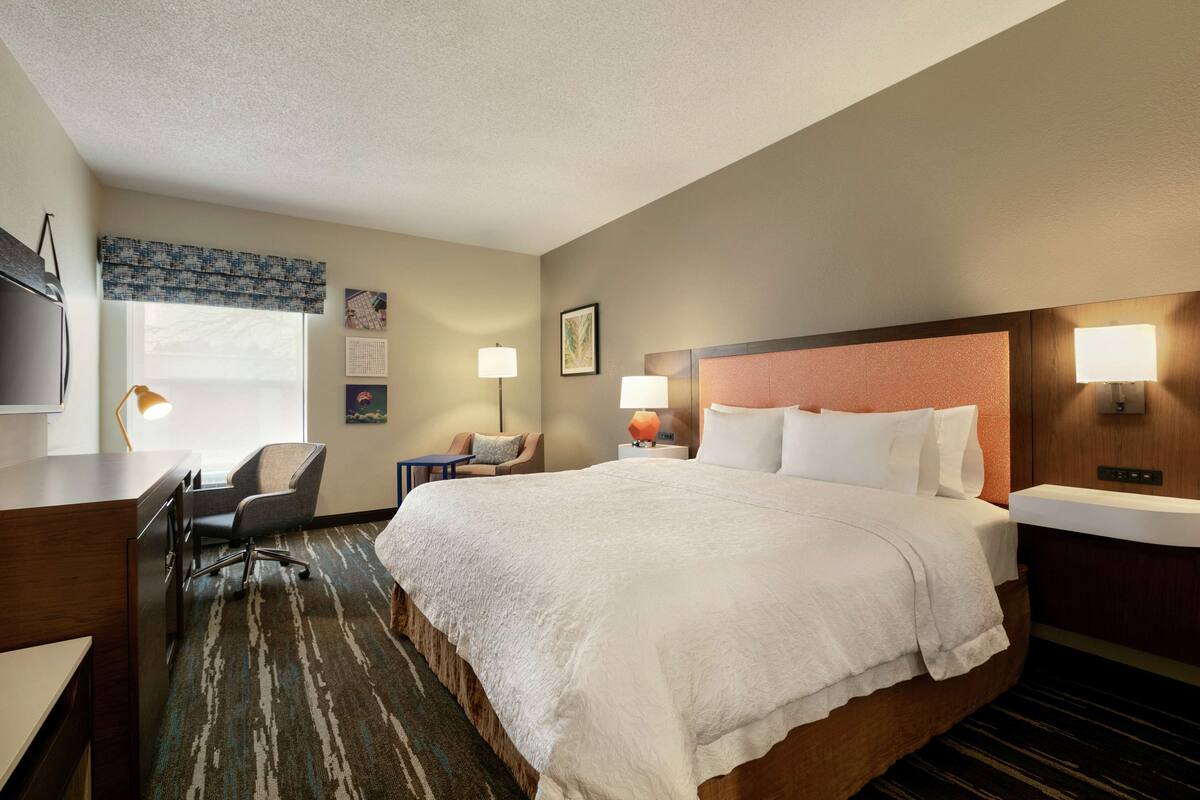 A hotel room with a large bed, a desk, a chair, a desk lamp, a wall-mounted TV, and a window with a view.