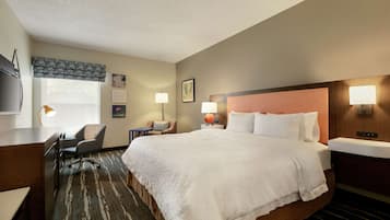 A hotel room with a large bed, a desk, a chair, a desk lamp, a wall-mounted TV, and a window with a view.