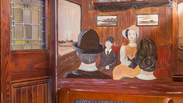 A vintage train carriage interior with wooden paneling, a window with a grid pattern, and a mural depicting a scene with three figures.