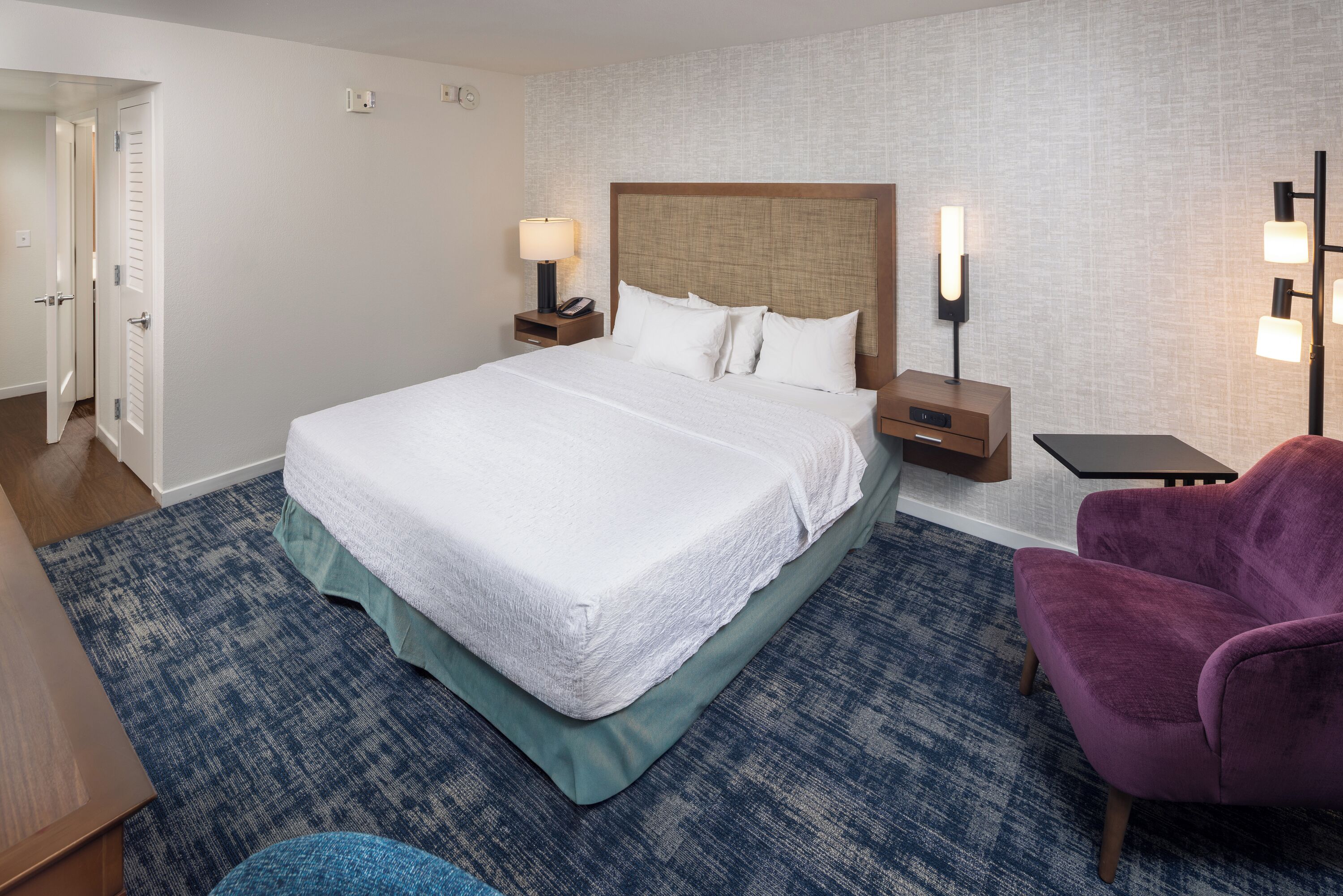 A hotel room with a large bed, two bedside tables with lamps, a purple armchair, and a blue carpet.