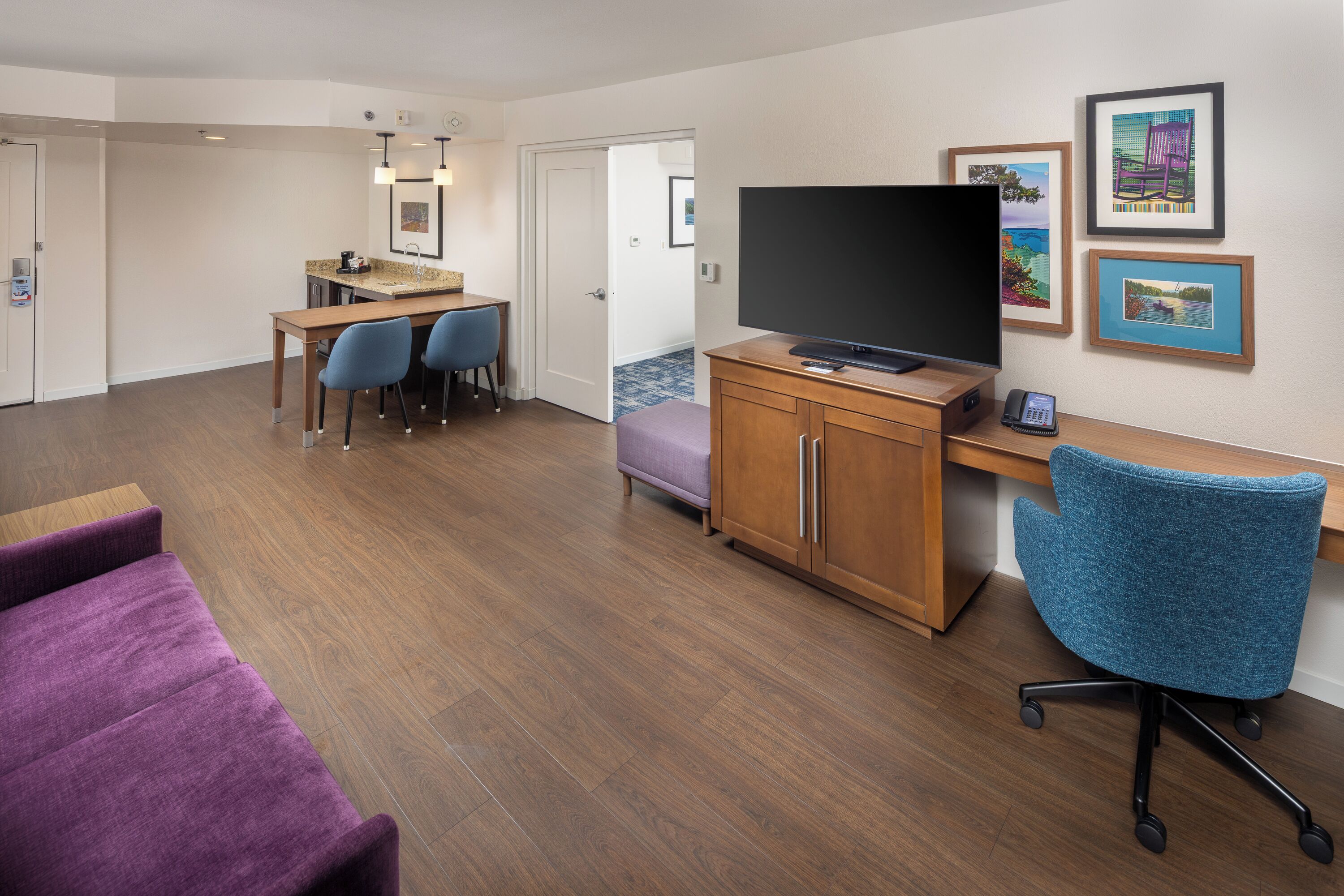 A modern hotel room with a wooden floor, a desk with a flat-screen TV, a blue office chair, a purple sofa, and a small kitchenette.