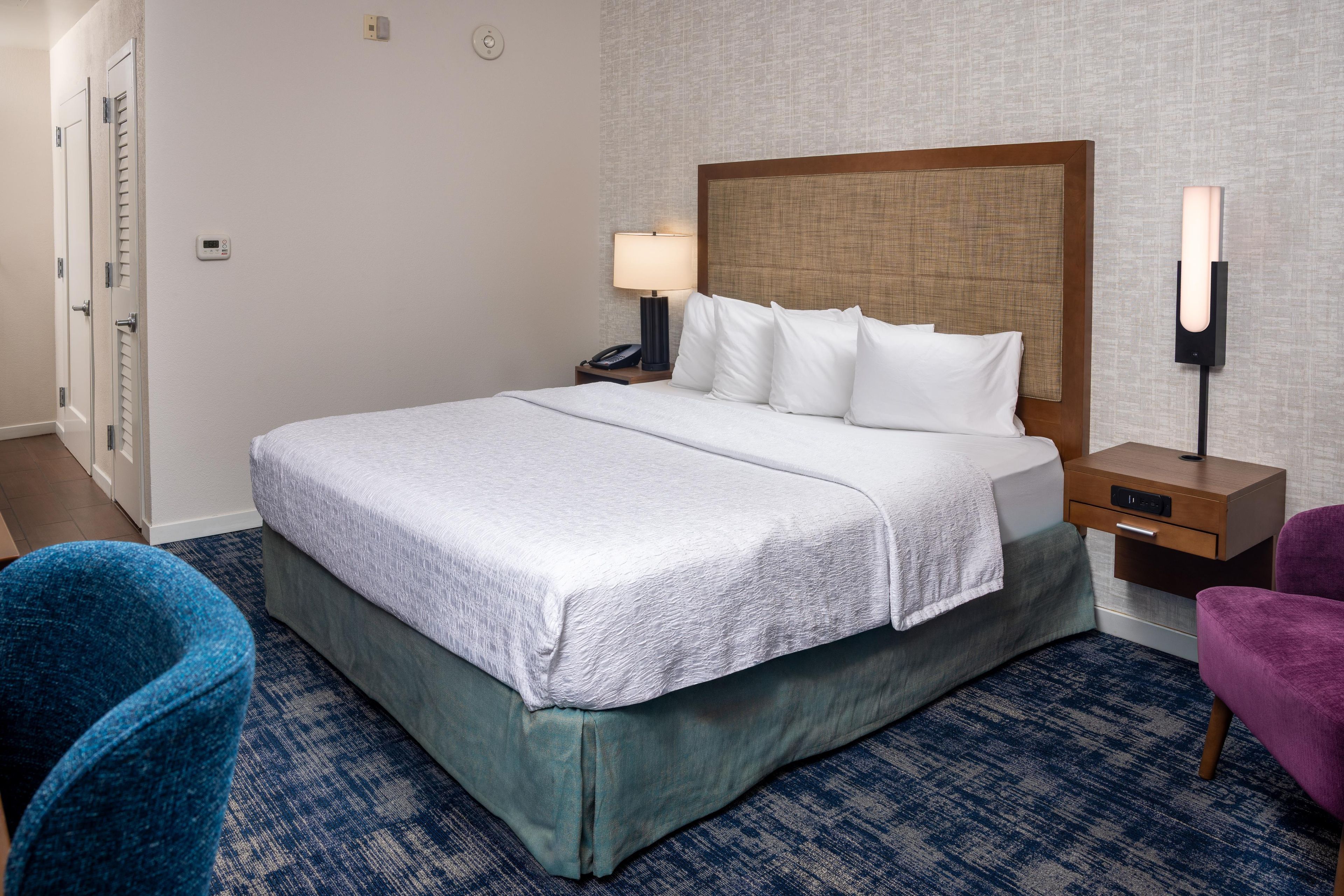 Habitación de hotel con una cama grande, una mesita de noche con lámpara, una silla y una luz en la pared.