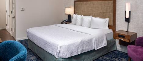 Habitación de hotel con una cama grande, una mesita de noche con lámpara, una silla y una luz en la pared.
