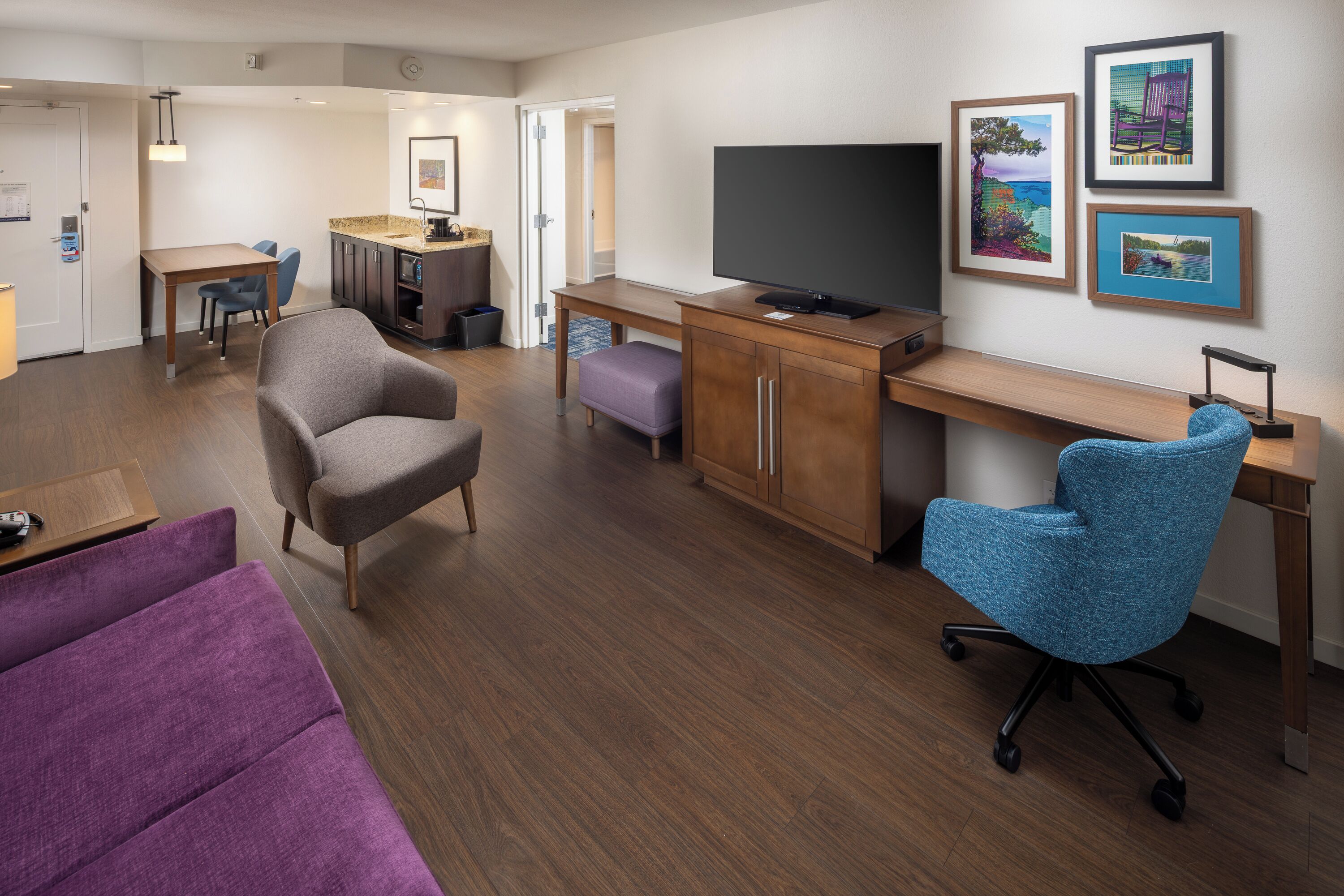 Una habitació d'hotel moderna amb escriptori de fusta, un gran televisor de pantalla plana, un butac armchair còmode, un sofà morat i una zona de menjador amb taula i cadires.