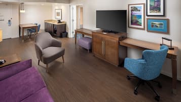 A modern hotel room with a wooden desk, a large flat-screen TV, a comfortable armchair, a purple sofa, and a dining area with a small table and chairs.
