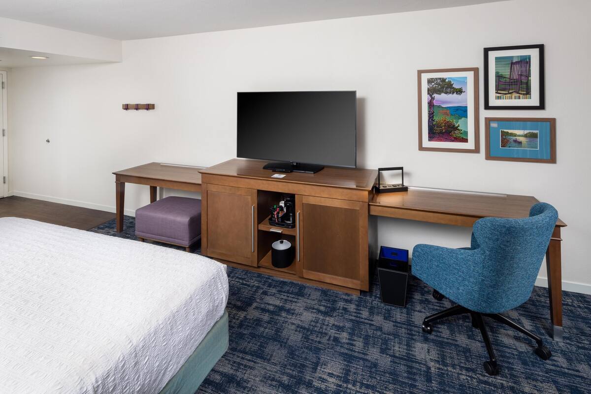 Una habitación de hotel con cama, escritorio con televisor, silla y cuadros en la pared.