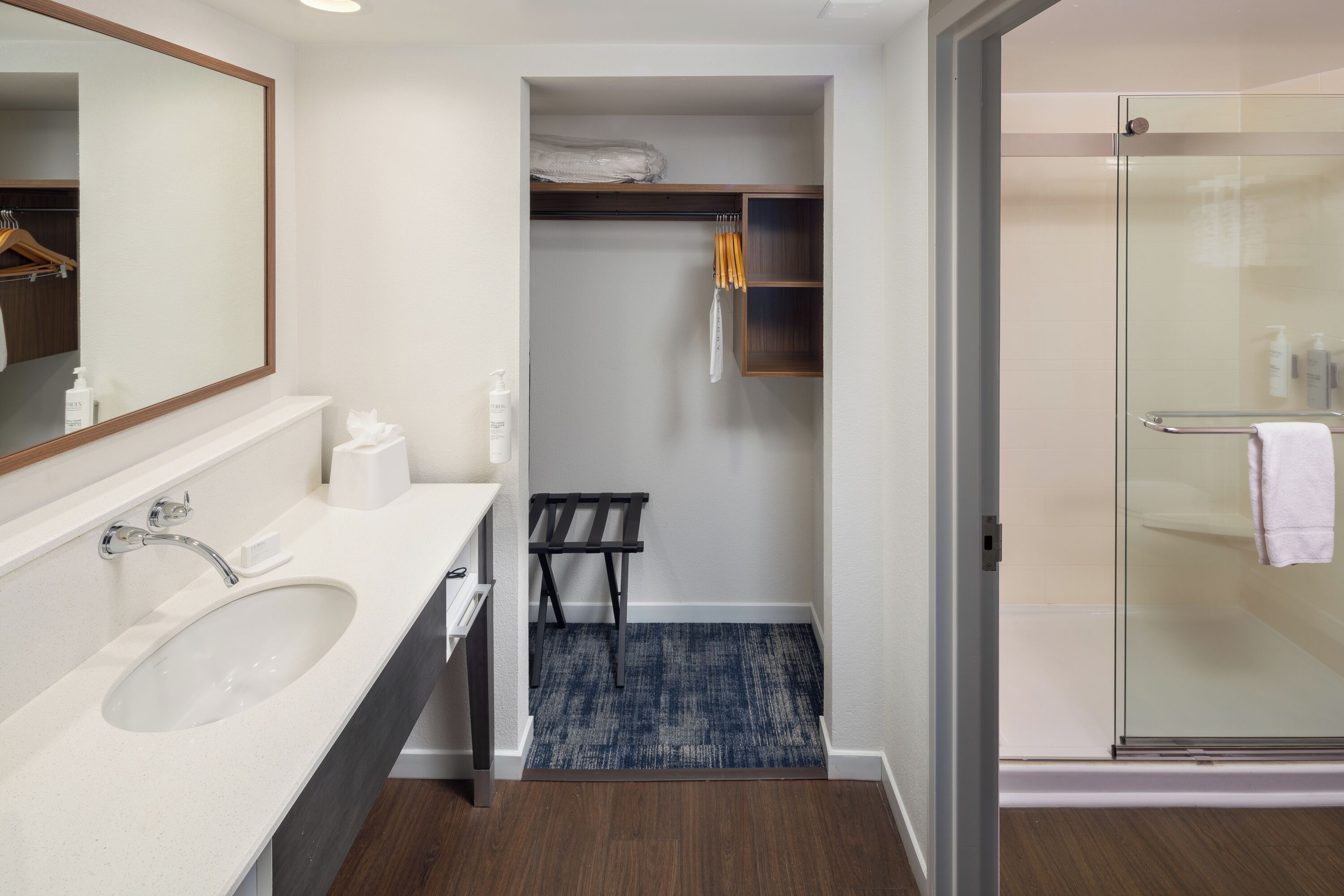 A modern bathroom with a large mirror, a white sink, a glass shower enclosure, and a walk-in closet with shelves.