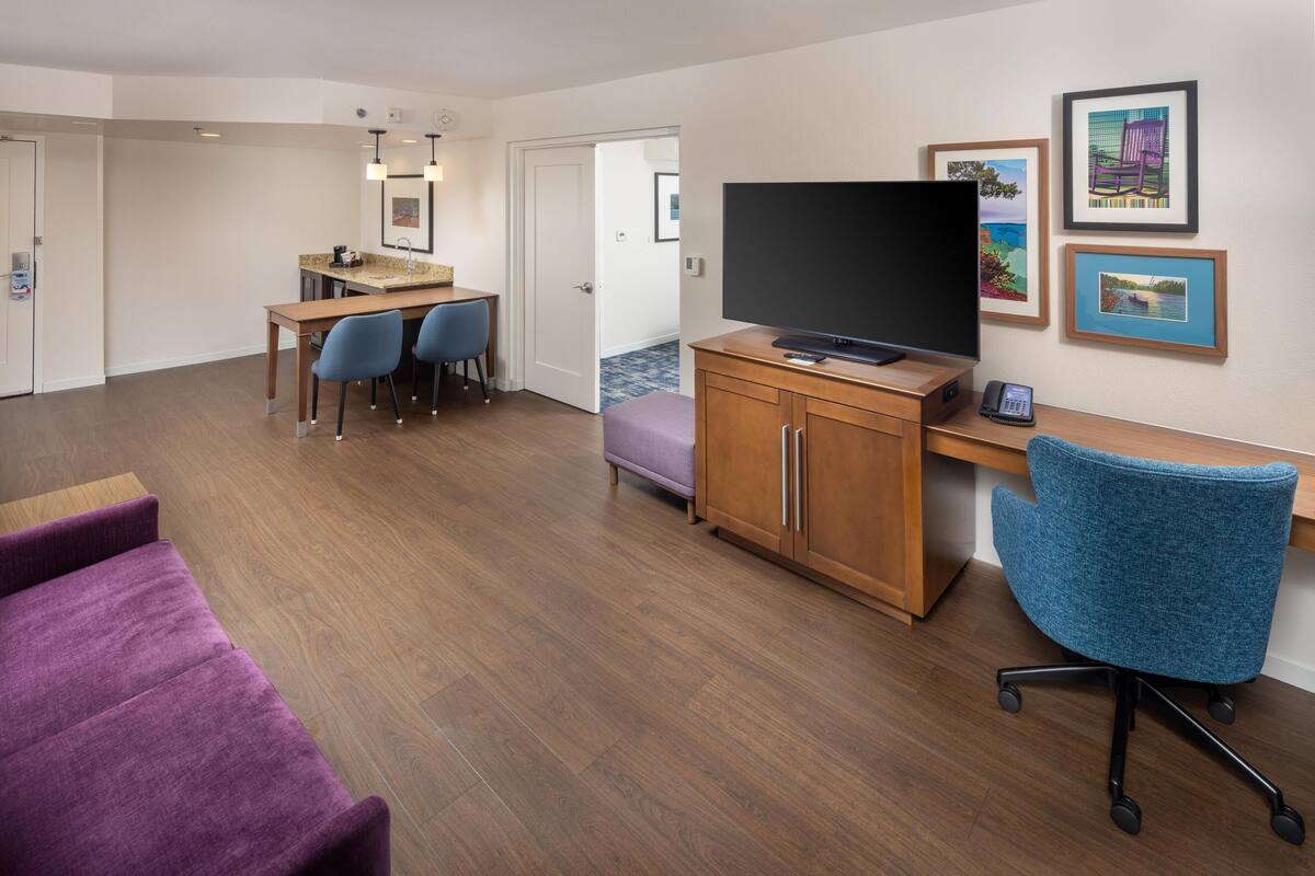 Habitación de hotel con piso de madera, un escritorio con televisor de pantalla plana, una silla de oficina azul, un sofá púrpura y una pequeña kitchenette.