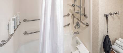 A bathroom with a bathtub, shower, and handrails. There are toiletries on a shelf and a towel rack with towels.
