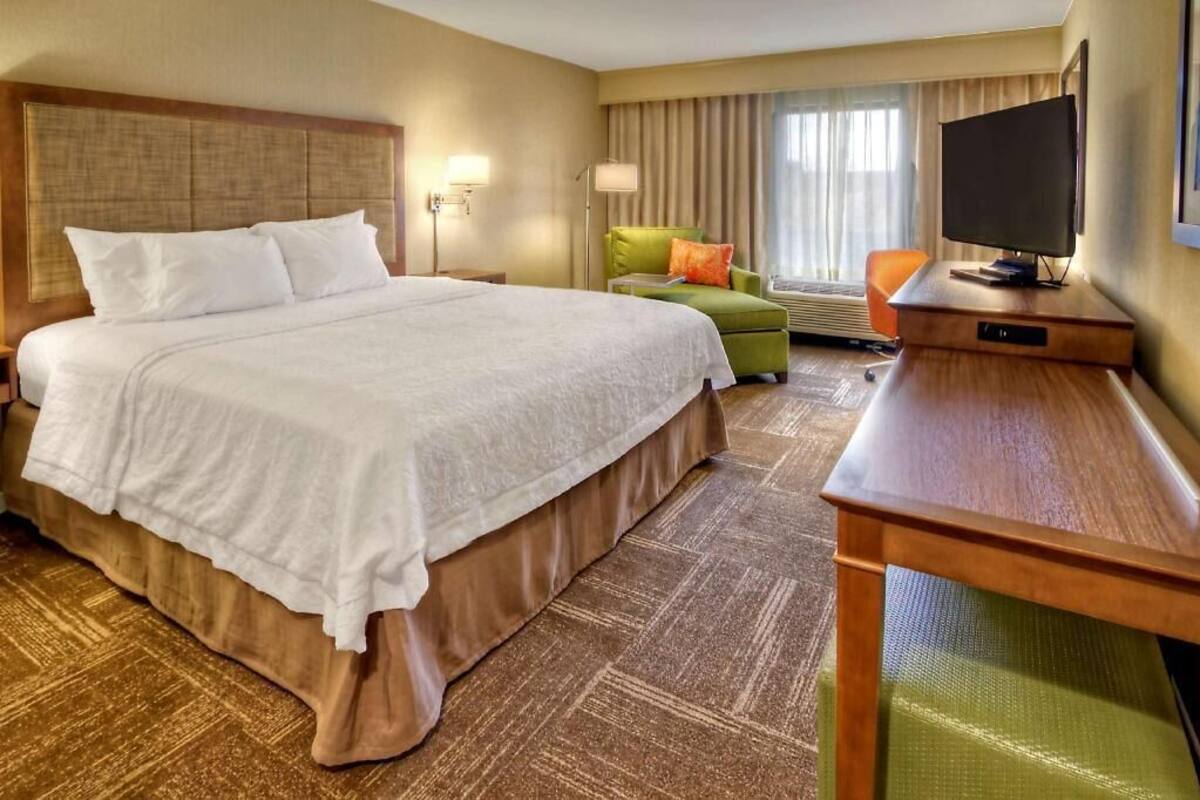 Habitación de hotel con una cama grande, un escritorio con televisor, una silla y un ventanal con cortinas.