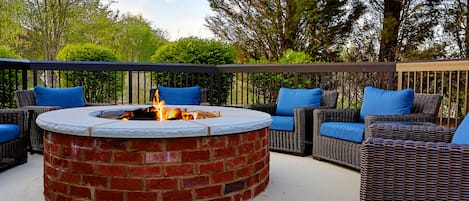 A circular brick fire pit with a white top surrounded by wicker furniture with blue cushions.