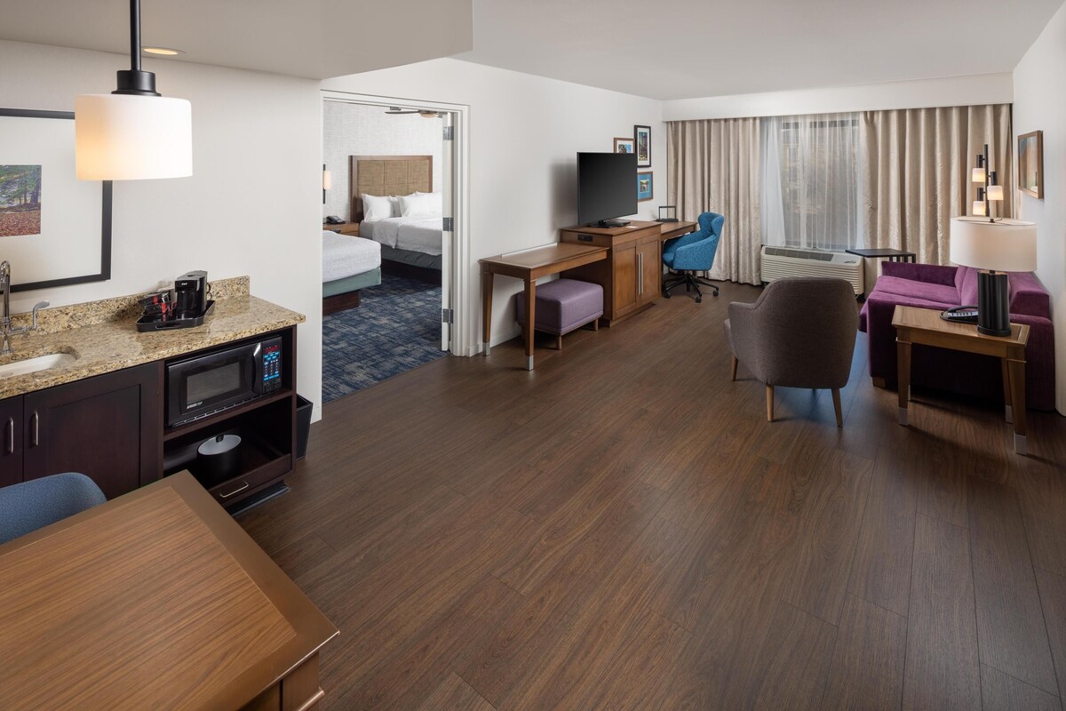 Une chambre d’hôtel équipée d’une kitchenette, d’un lit, d’un bureau avec un ordinateur, d’une télévision et d’un coin salon comprenant une chaise et un canapé.