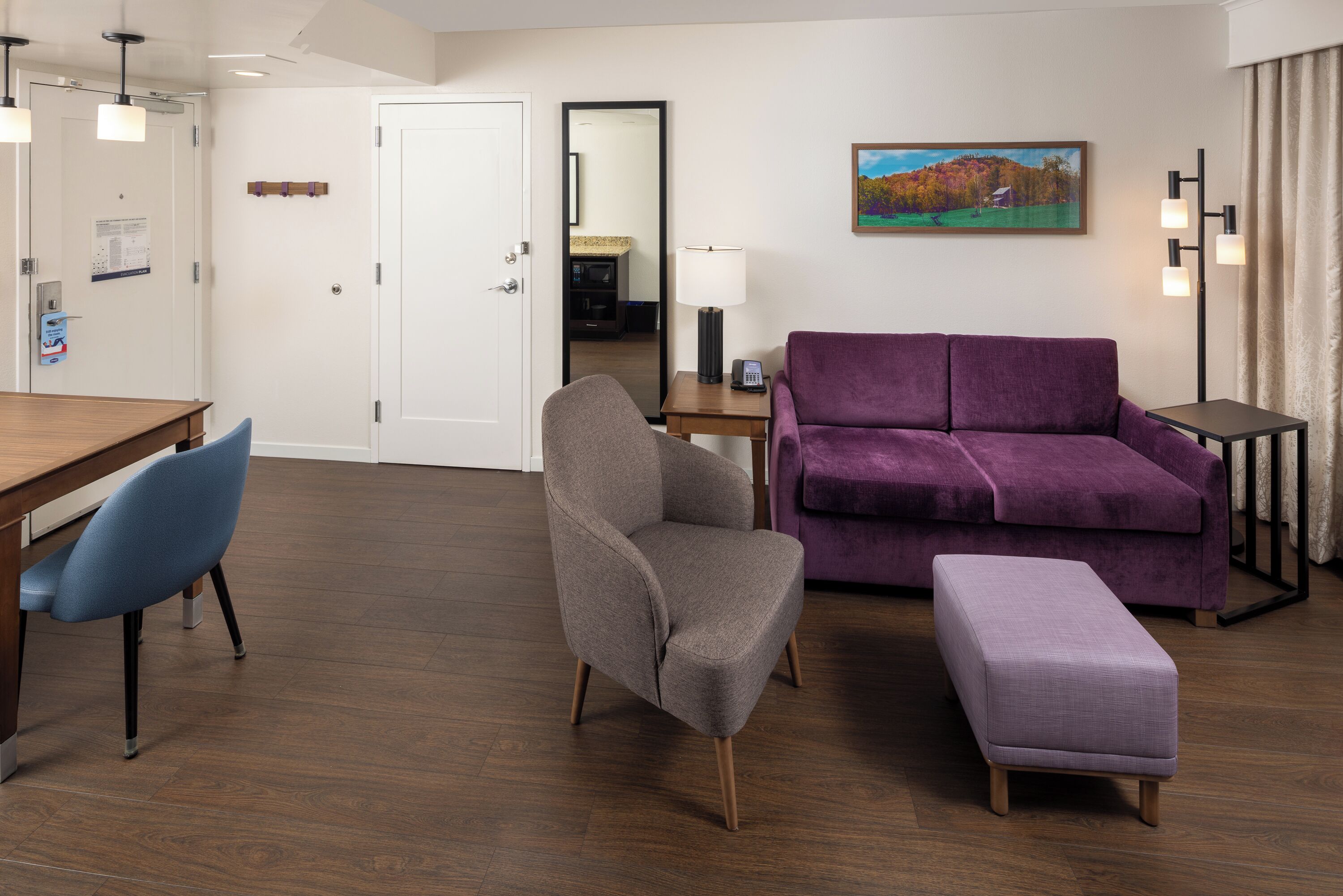 A modern hotel room with a wooden dining table, two chairs, a purple sofa, a matching ottoman, a small side table with a lamp, and a painting on the wall.