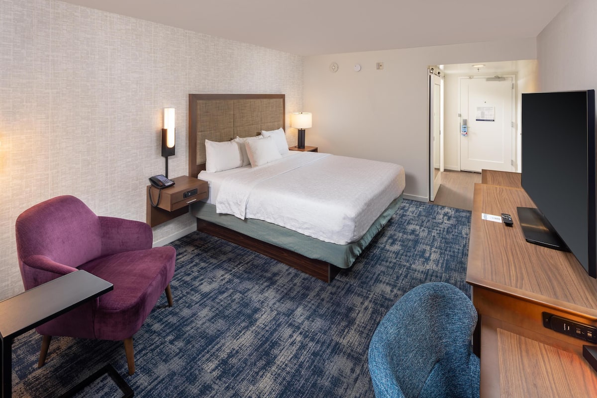 Habitación de hotel con cama, dos mesitas de noche, televisor, escritorio, silla y piso alfombrado.