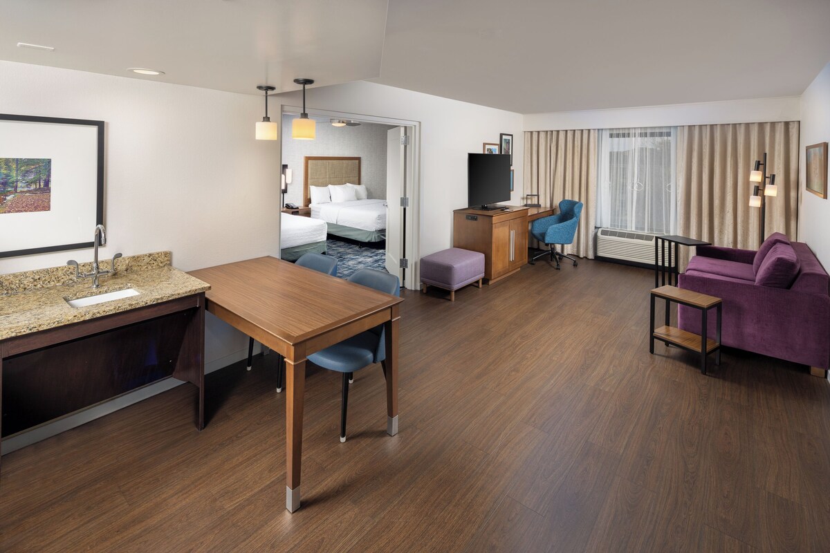 Une chambre d’hôtel avec un bureau en bois, un lit, une télévision, un canapé violet et une vue sur l’extérieur par la fenêtre.
