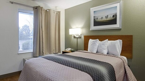 A hotel room with a large bed, a nightstand with a lamp, a framed picture on the wall, and a window with curtains. - Suburban Studios Winston Salem-University Parkway (Winston-Salem)